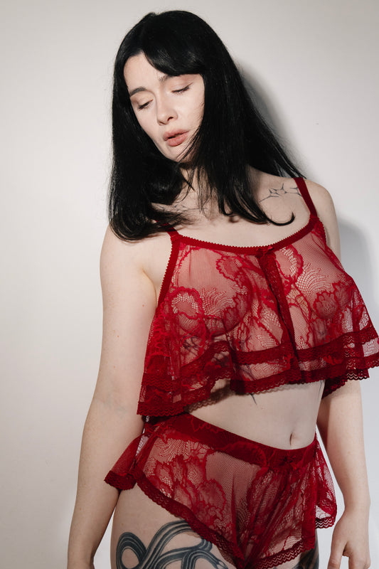 A woman modeling a red sheer lace lingerie set against a plain light gray wall. The set includes a cropped camisole top and matching high-waisted briefs.