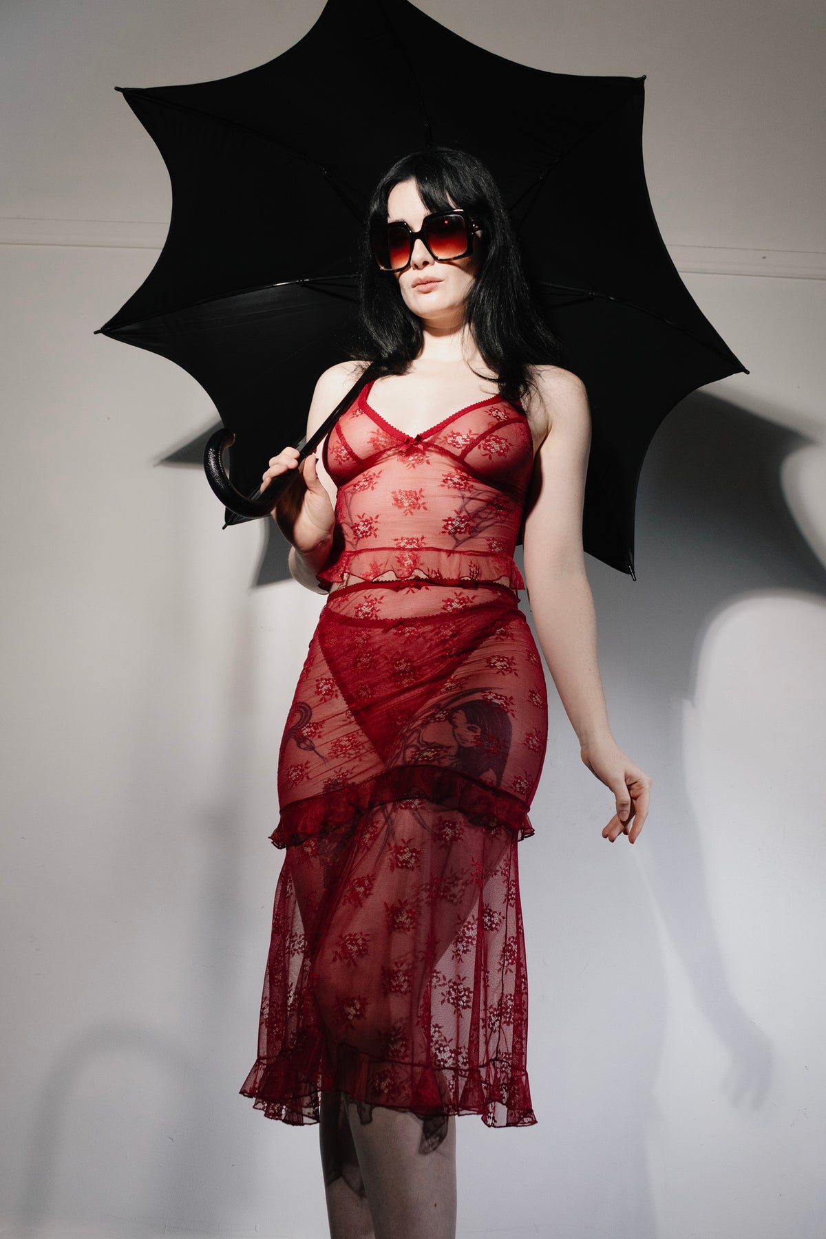A model wearing a sheer red lace dress with floral patterns and holding a black umbrella, posing against a white studio background.