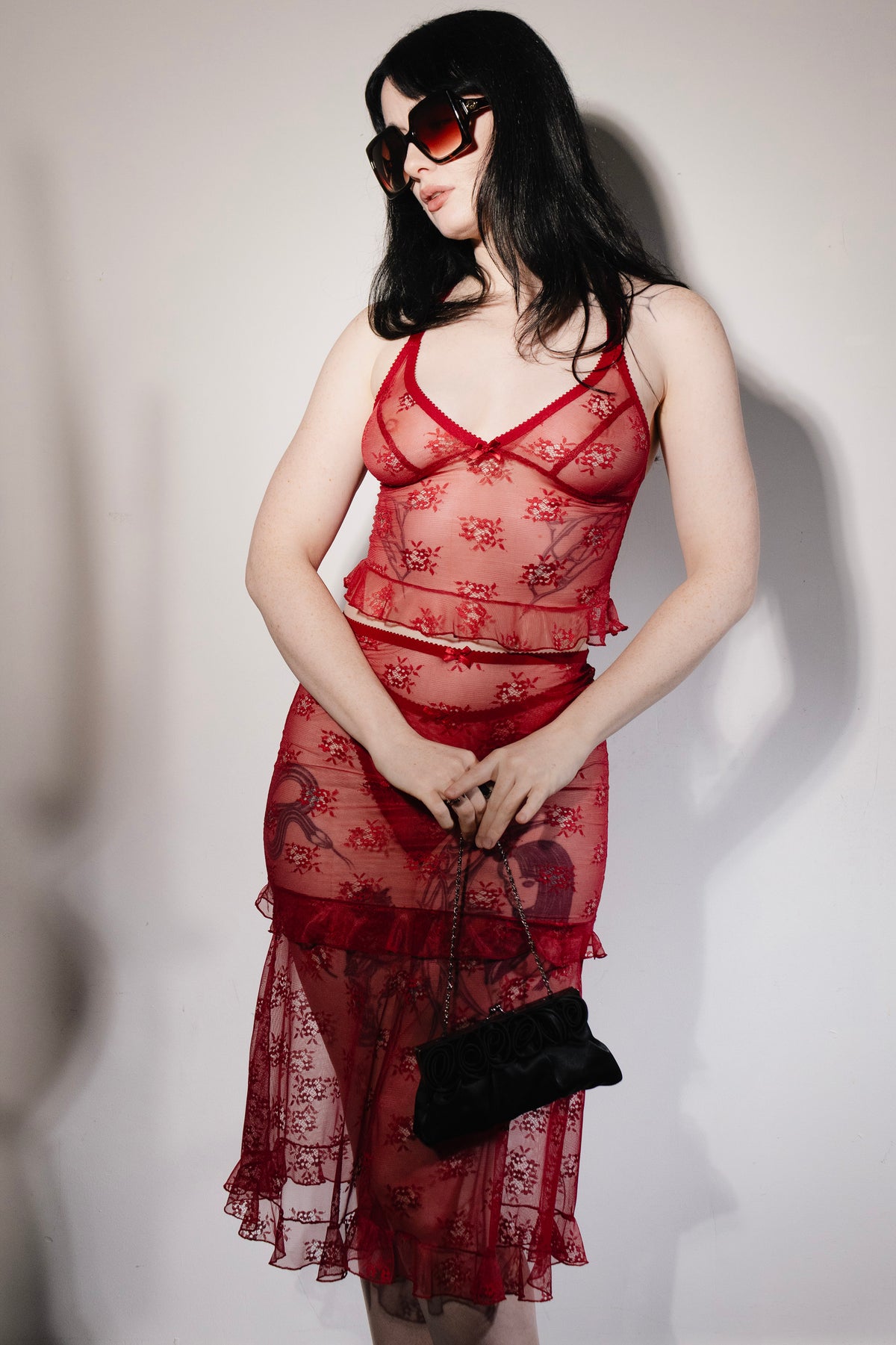 A model wearing a sheer red lace two-piece lingerie set with a floral pattern, featuring a camisole top and a ruffled midi skirt, accessorized with a black handbag and dark sunglasses against a plain light background.