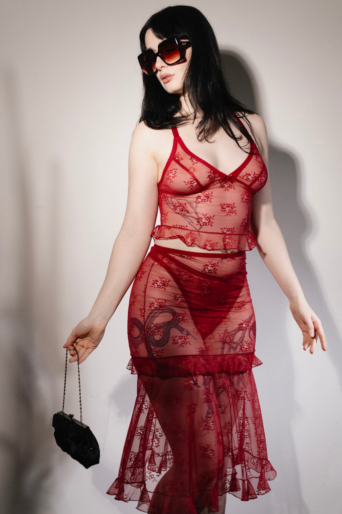 A woman modeling a sheer red lace two-piece lingerie set with a crop top and ruffled skirt, accessorized with dark sunglasses and a small black handbag against a plain light background.