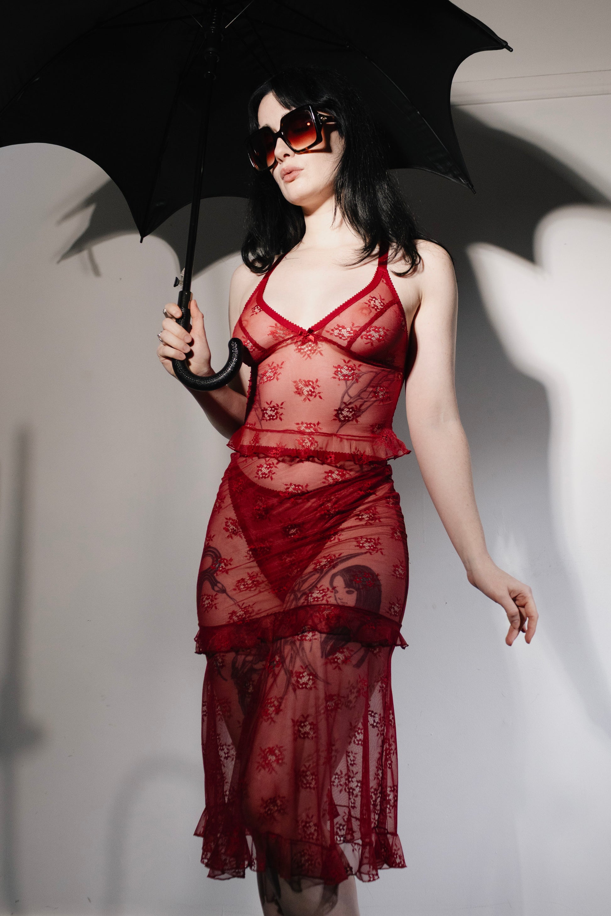 A model wearing a sheer red lace dress with floral embroidery, holding a black umbrella against a white studio background.