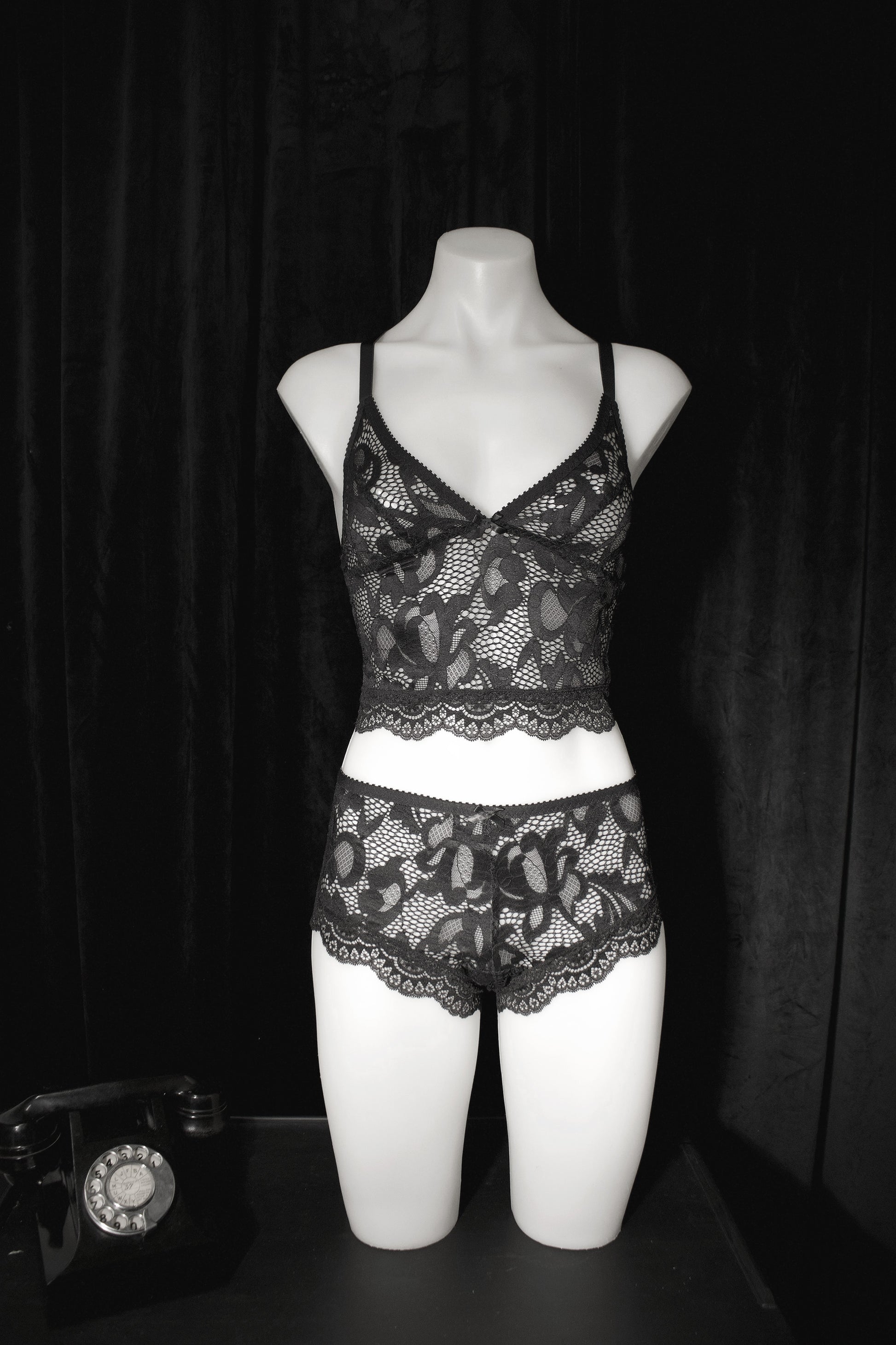 Black lace lingerie set with a camisole top and matching briefs displayed on a white mannequin torso against a black curtain backdrop.