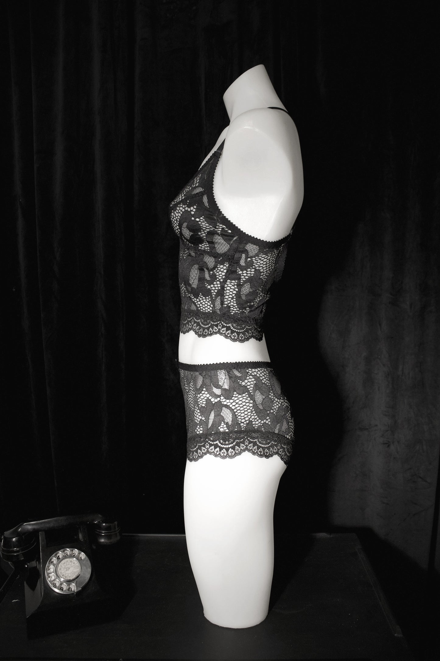 Black lace lingerie set displayed on a white mannequin torso against a dark curtain background, with a vintage rotary telephone visible at the lower left.