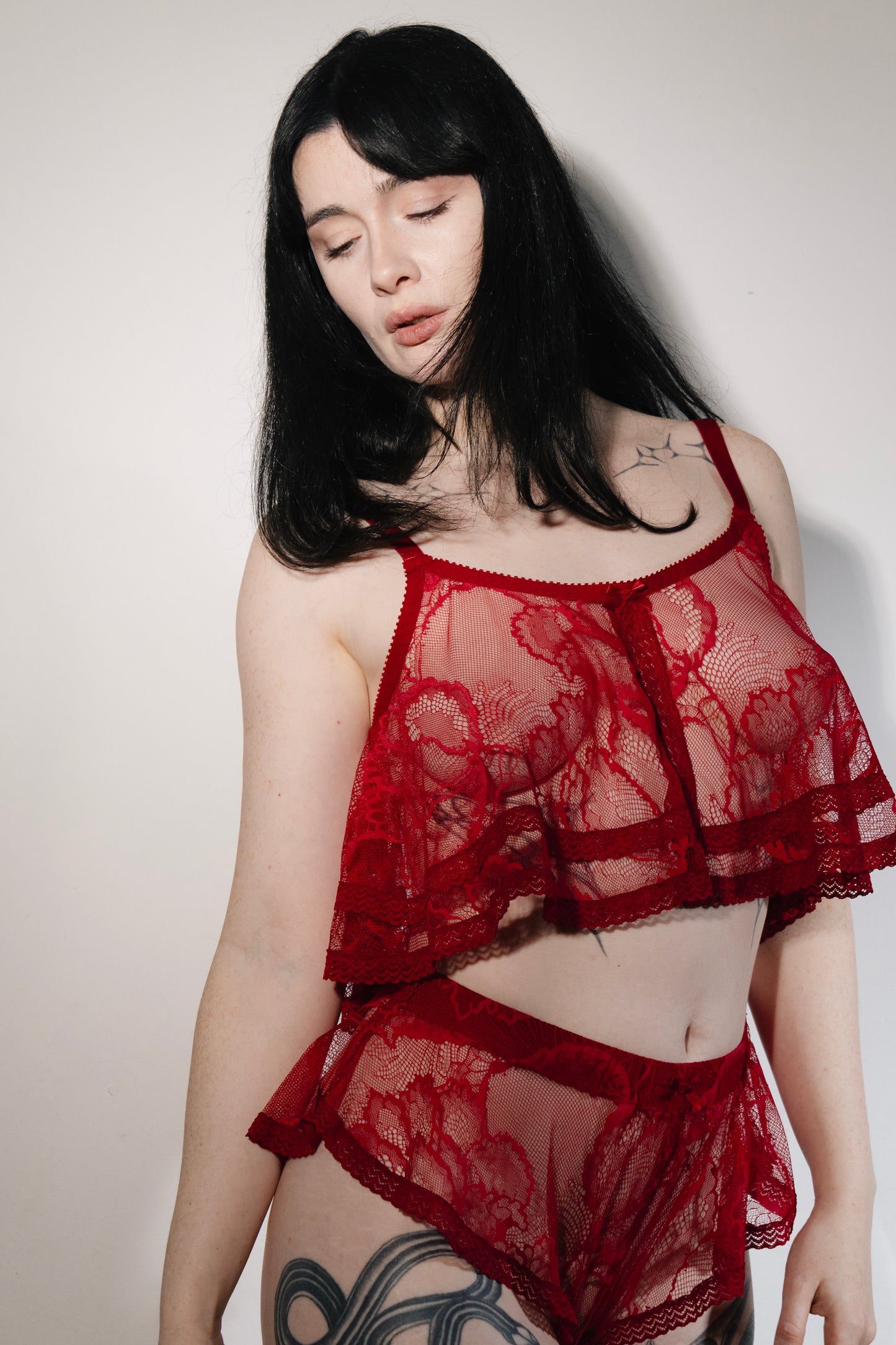 A woman modeling a red sheer lace lingerie set against a plain light gray wall. The set includes a cropped camisole top and matching high-waisted briefs.