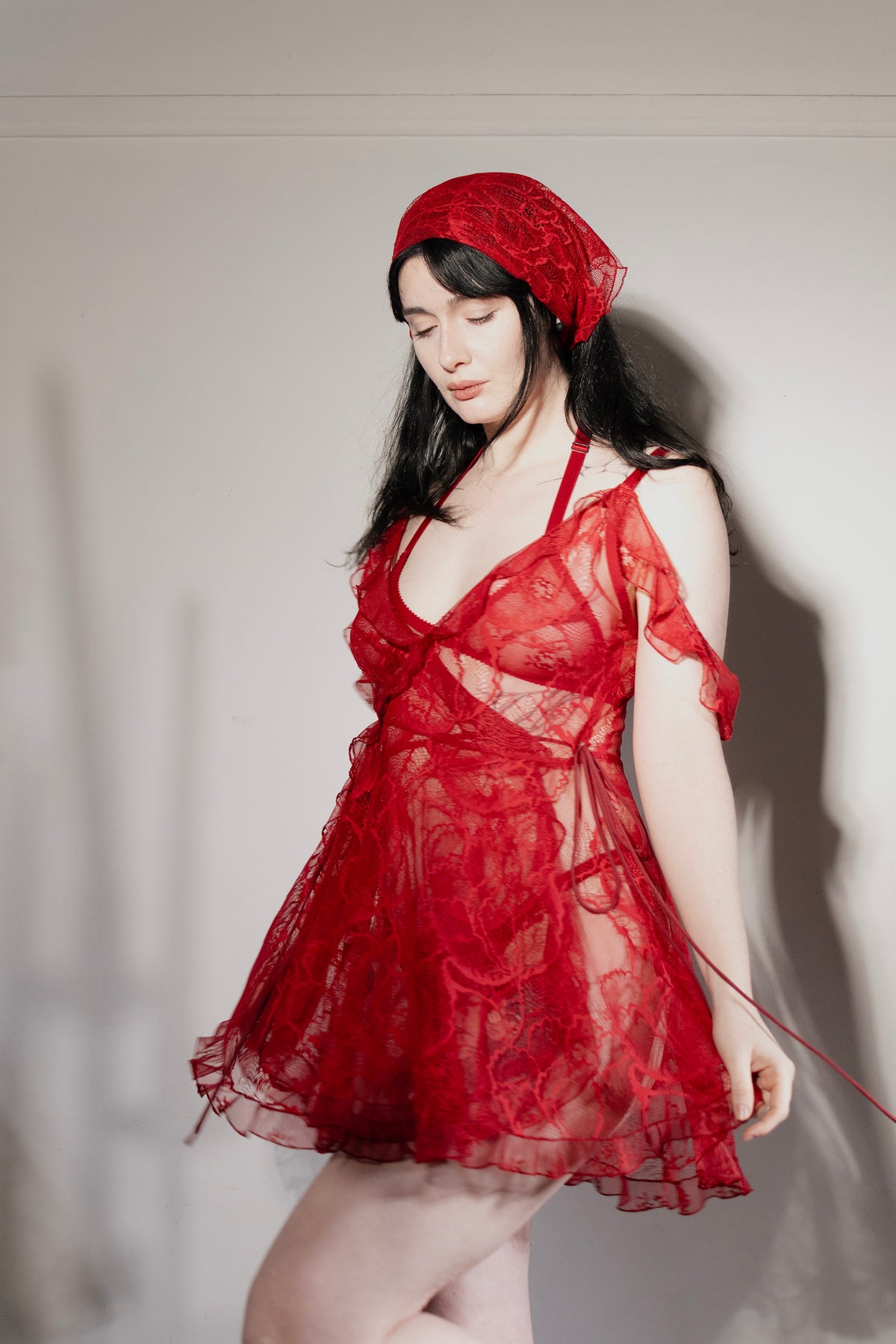 A woman wearing a sheer red lace mini dress with a matching red lace headscarf, posed against a plain light gray wall.